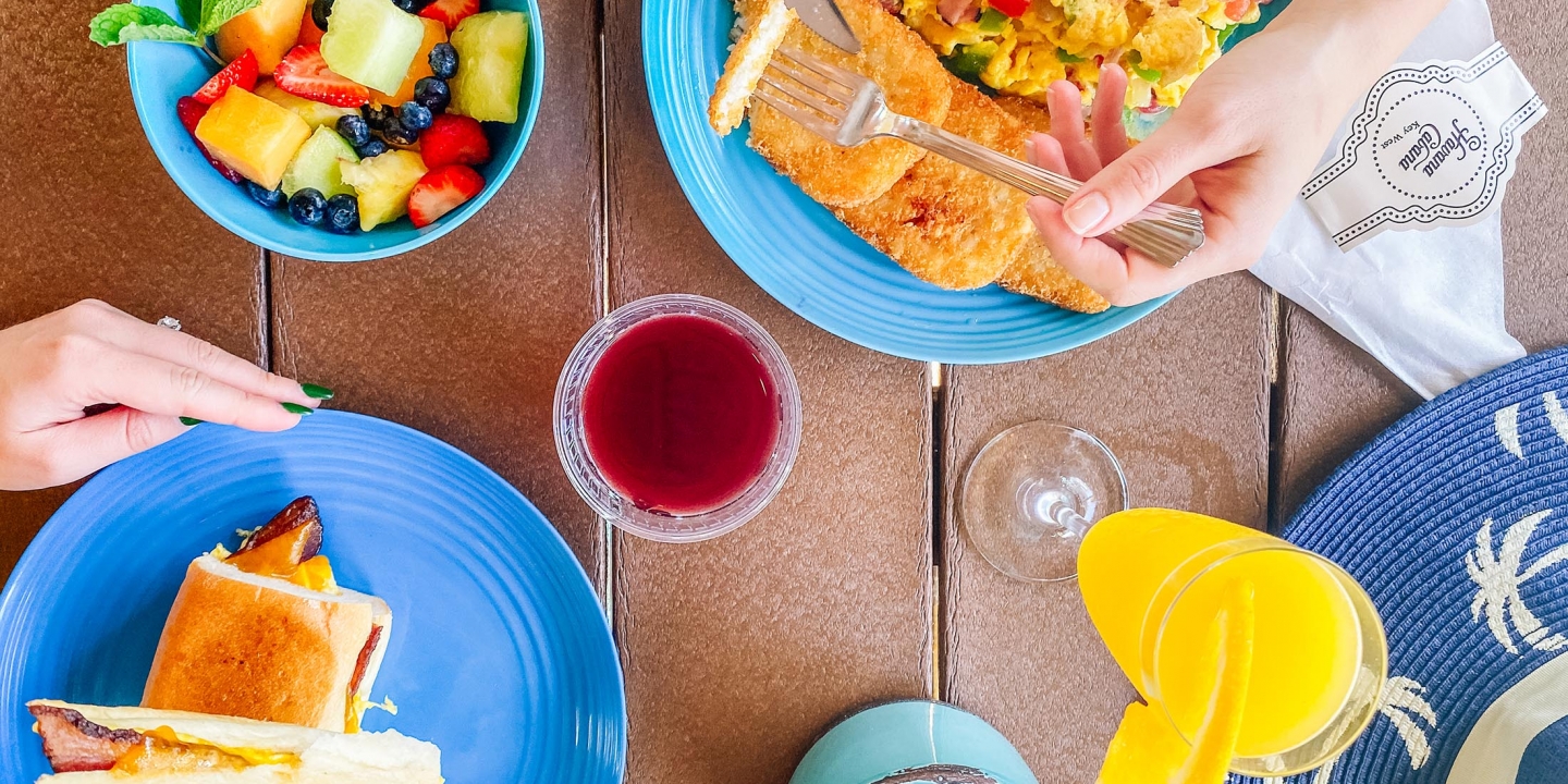 A hand holding a sandwich near a fruit bowl, while another pair of hands is using cutleries to cut chops next to scrambled egg