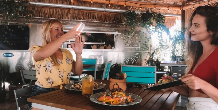 A woman photographing of her friend posing with her lunch at the Tiki Hut