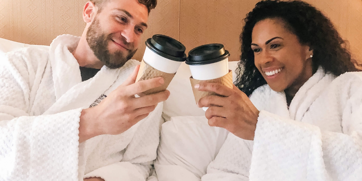 A couple in robes sitting on a bed and clinking their disposable coffee cups